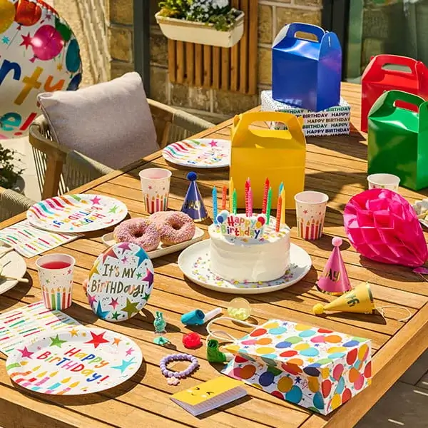 Colourful birthday party table with a cake, party hats, gift bags, confetti, snacks, and balloon-print tableware.
